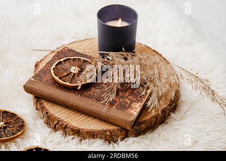 Vintage-Papierbuch auf Pinienholz-Scheibe mit trockenem Schilf und getrockneten Orangenscheiben. Aroma-Kerzen brennen, Gegenstände auf Schaffell. Gemütliches Herbstset im Winter. Stockfoto