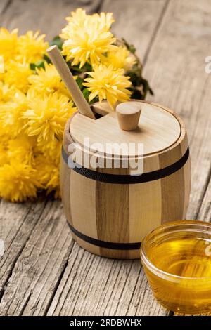 Honig in einem kleinen Holzfass und einer transparenten Glasschüssel, einem Holzdipper, gelben Blumen auf einem gereiften Holzhintergrund, Nahaufnahme. Stockfoto