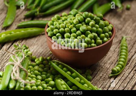 Frische organische grüne Erbsen in geschlossenen und offenen Schoten, verstreute Erbsensamen, geschälte grüne Erbsen in einer Tonschüssel auf einem gereiften Holzhintergrund. Stockfoto