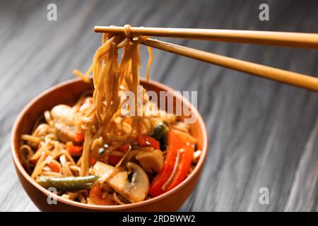 Teller mit Buchweizennudeln mit Gemüse, Pilzen, Hühnerfleisch auf dunklem Hintergrund. Chinesische Stöcke nehmen japanische Soba aus einer Tonschüssel Stockfoto