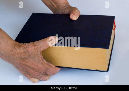 Eine alte Hand hält ein altes dickes Buch auf einer weißen Oberfläche, über der Aussicht Stockfoto