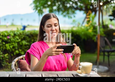 Wunderschöne brünette Frau, die am Tisch sitzt und ein Foto von ihrem Cappuccino macht Stockfoto