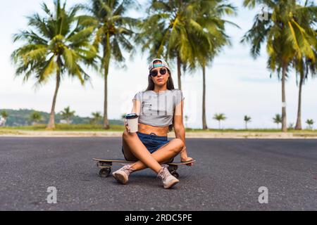 Junge, schöne Frau mit Mütze, die auf einem Skateboard sitzt und Kaffee trinkt Stockfoto