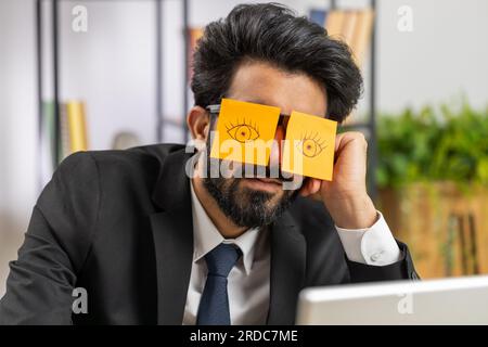 Ineffizienter, müder, junger Geschäftsmann, der auf einem Laptop mit Augenaufklebern auf dem Gesicht am Schreibtisch im Büro schläft. Indischer fauler Manager, Freiberufler. Geschäftsleute schummeln in den Schlaf Stockfoto