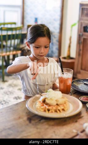 Kleines asiatisches Mädchen, das süße belgische Waffeln mit Banane und Sahne in einem Café isst Stockfoto