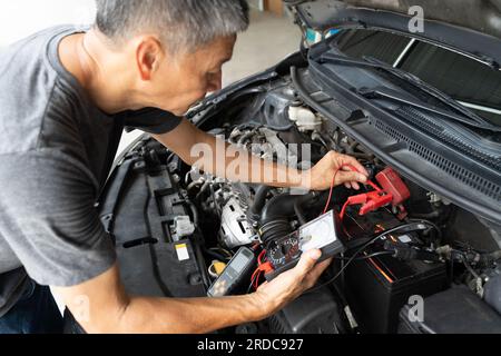 Ein asiatischer Mechaniker mittleren Alters, der mit einem Batterieprüfer den Status der Autobatterie überprüft, mit selektivem Fokus auf die Hände Stockfoto