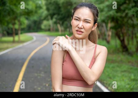 Junge, atraktive Asiatin in Fitnesskleidung, die ihre linke Hand über ihre schmerzhafte rechte Schulter legt, während sie auf einer Laufstrecke eines örtlichen Parks steht Stockfoto