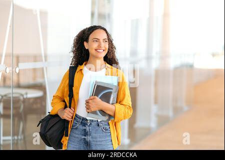 Bildungskonzept, weibliche Studentin. Glückliche, reizende Studentin mit gemischter Rasse, mit einem Rucksack, mit Büchern und Notizbüchern in der Hand, steht neben dem Universitätscampus, sieht weg und lächelt. Kopierraum Stockfoto