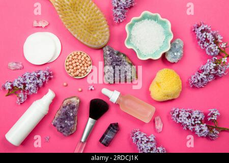 Von oben sehen Sie verschiedene Behälter mit weißen Kosmetik- und Schönheitsprodukten mit Fliederblüten (Syringa) und halbkostbaren Amethyst-Kristallen. Stockfoto