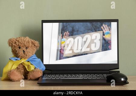 Ein Foto eines kleinen Mädchens auf einem Laptop-Bildschirm mit dem Text 2023, und die Flagge der Ukraine wird auf ihre Hände gezeichnet. Ansicht aus dem Fenster von Stockfoto