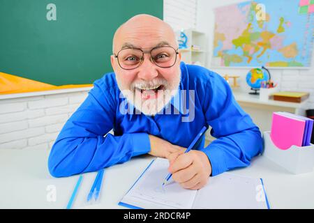 Glücklicher männlicher Trainer, Lehrer oder Universitätsprofessor, der im Klassenzimmer am Schreibtisch sitzt. Lehrertag. Der Lehrer bereitet den Unterricht im Klassenzimmer vor. Lernen Stockfoto