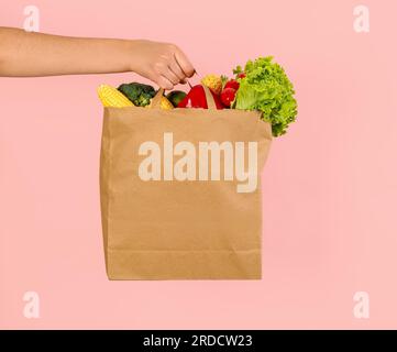 Weibliche Hand mit Papiertüte voller frischem Gemüse auf pinkfarbenem Hintergrund Stockfoto