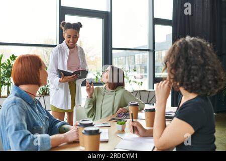 Positives afroamerikanisches Sofa liest vom Notebook zu multiethnischen weiblichen Mitgliedern, die in der Nähe von Kaffee zum Mitnehmen sitzen, und Smartphones im Interessenverein, Mutual Stockfoto
