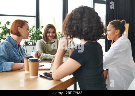 Fröhliche junge Frau, die mit einer multiethnischen weiblichen Gruppe in der Nähe von Smartphones und Kaffee spricht, um in eine freundliche Atmosphäre von Interessenverein, gegenseitiger Unterstützung und zu gehen Stockfoto