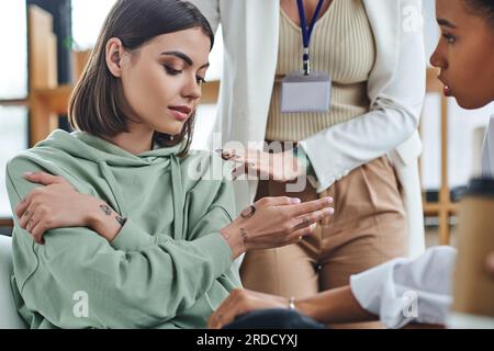 Professionelle Psychologin beruhigende, enttäuschte tätowierte Frau, die neben einer afroamerikanischen Freundin auf verschwommenem Vordergrund in der Sprechstunde sitzt, EM Stockfoto