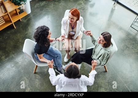 Aus einem großen Blickwinkel betrachten wir freudige, multiethnische weibliche Freunde und rothaarige Motivationslehrerin, die im Kreis sitzt und Händchen hält, während sie über Gruppentherapie spricht. Stockfoto