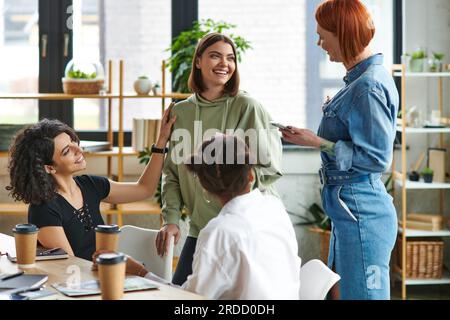 Optimistische, multikulturelle weibliche Freunde, die sich mit einer zufriedenen jungen Frau unterhalten, während sie sich in der Nähe des Tisches mit Pappbechern und Kaffee unterhalten, um sie zu interessieren C. Stockfoto