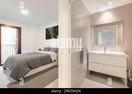 Blick von einem gemütlichen Bad in stilvollem Beige mit Waschbecken mit Schrank und Spiegel auf das Schlafzimmer mit einem Bett in einem hellen Zimmer. Stilvoll Stockfoto