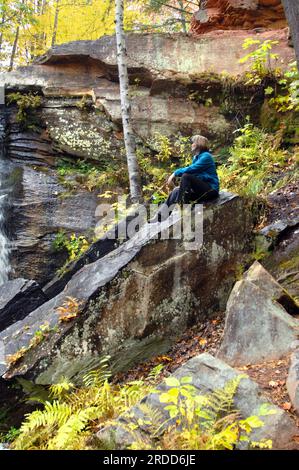 Neben den ungarischen Fällen auf der oberen Halbinsel, Michigan, sitzt eine Frau auf einem großen Felsen. Hintergrund und Blätter in gelben Herbstfarben. Stockfoto