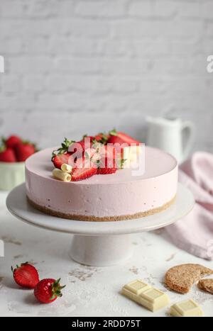 Erdbeerkäsekuchen auf einem weißen Kuchenstand, dekoriert mit frischen Erdbeeren und weißer Schokolade. Rosa Serviette und weißer Milchbehälter. Stockfoto