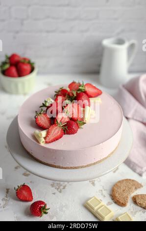 Erdbeerkäsekuchen auf einem weißen Kuchenstand, dekoriert mit frischen Erdbeeren und weißer Schokolade. Rosa Serviette und weißer Milchbehälter. Stockfoto