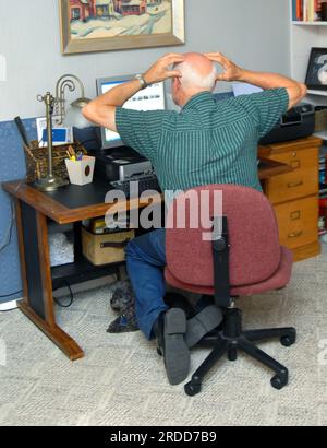 Ein erwachsener Mann macht sich Sorgen über die Ergebnisse auf dem Computerbildschirm. Er ist aufgebracht und hält seinen Kopf in den Händen. Er hat ein Privatunternehmen, komplett mit Schreibtisch. Stockfoto
