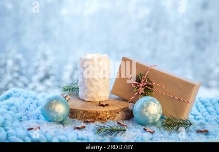 Hintergrundinformationen zu Weihnachtsprodukten. Kerze mit weißem Schneeballmuster brennt, Weihnachtsgeschenk verpackt in braunes Papier mit weiß-roter Schnur. Stockfoto