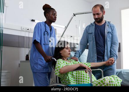 afroamerikanische Krankenschwester, die schwangere Frau im Rollstuhl in den Entbindungsraum bringt, um das Baby zur Geburt zu bringen. Patientin mit Schwangerschaft, die sich auf die Geburt vorbereitet, während sie durch den Ehemann auf der Krankenstation getröstet wird Stockfoto