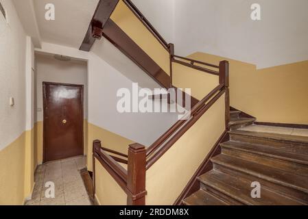 Die alten Treppen mit braunen Holzgeländern und Stufen und dunkelcremefarbenen und weißen Wänden führen zu einer Wohnung mit Tür der gleichen Farbe Stockfoto