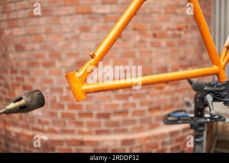 Eine nicht wiedererkennbare Person, die mit einem Lötbrenner die Farbe eines orangefarbenen Fahrradrahmens entfernt, der gerade in seiner Werkstatt renoviert wird. Stockfoto