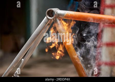 Manuelle Fahrradrenovierungsarbeiten, Lackentfernung mit einem Brennerfeuer, um die Farbe eines orangefarbenen Fahrradrahmens zu ändern. Nahkomposition ohne Erbsen Stockfoto