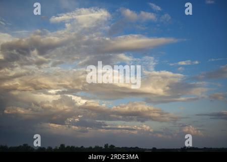 Blauer Himmel mit weichen weißen Kumuluswolken, erleuchtet von der Sonne, die in der Ferne untergeht, und wirft einen weichen Orange- und Pink-Farbton über das Land Stockfoto