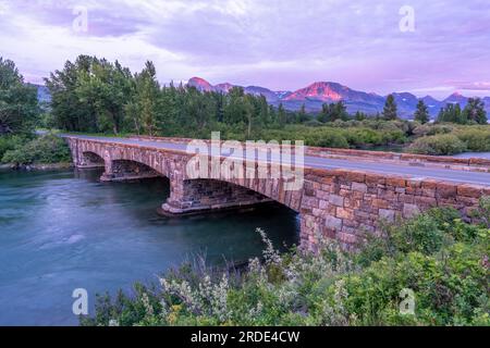 Eine Steinbrücke, die einen breiten Fluss mit Bäumen im Hintergrund im sanften Licht vor Sonnenaufgang überspannt, Saint Mary Lake Bridge, Glacier National Stockfoto