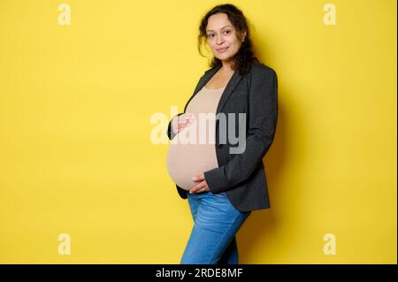 Attraktive lateinamerikanische Schwangere in stilvollem Blazer, Body und Blue Jeans, lächelt süß in die Kamera, posiert mit den Händen am Bauch Stockfoto