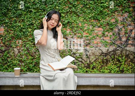 Eine entspannte und glückliche junge asiatische Frau in lässiger Kleidung hört ihre Musik auf ihren Kopfhörern, während sie sich auf einer Bank im Stadtpark entspannt. Stockfoto