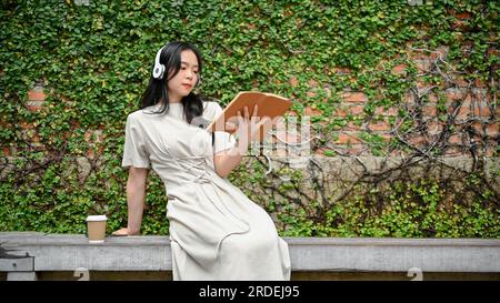 Eine charmante und entspannte junge Asiatin in einem schönen Kleid liest ein Buch und hört Musik auf ihren Kopfhörern, während sie auf einer Bank in einem sitzt Stockfoto