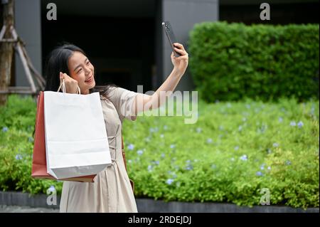 Eine schöne und glückliche junge Asiatin in lässiger Kleidung macht Selfies mit ihren Einkaufstaschen, während sie auf der Straße in der Stadt spaziert. Urbane Außenseite Stockfoto