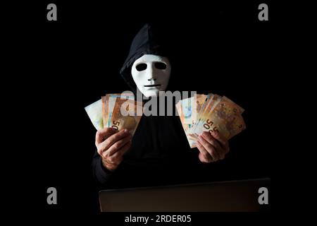 Schwarz gekleideter Hackermann mit weißer Maske und schwarzer Kapuze, der Euro-Banknoten in den Händen hält, Cyberkriminalität, isoliert auf schwarzem Hintergrund Stockfoto