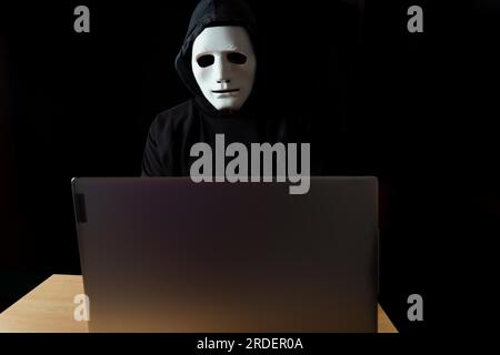Männlicher Hacker, schwarz gekleidet, mit einer Kapuze und einer weißen Maske hinter einem Computer Stockfoto