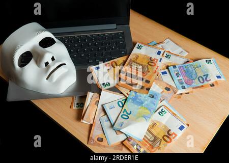 Auf einem Laptop befinden sich eine weiße Maske und ein Haufen Euro-Banknoten, das Konzept der Cyberkriminalität Stockfoto