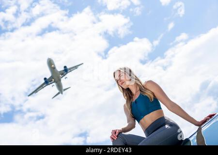 Ein schönes blondes Modell genießt einen Sommertag, während ein kommerzielles Flugzeug über dem Himmel fliegt Stockfoto