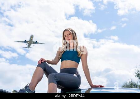 Ein schönes blondes Modell genießt einen Sommertag, während ein kommerzielles Flugzeug über dem Himmel fliegt Stockfoto