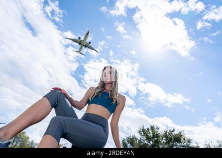 Ein schönes blondes Modell genießt einen Sommertag, während ein kommerzielles Flugzeug über dem Himmel fliegt Stockfoto