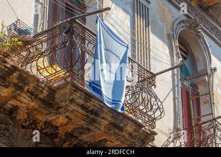 Havanna, Kuba - 27. Mai 2023: Ein blauer Turm, der im Wind trocknet. Der Textilgegenstand hängt von einem Balkon in einer verwitterten alten Gebäudefassade. Stockfoto