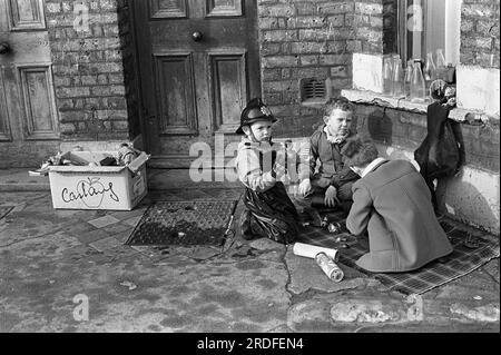 Kinder der 1970er Jahre spielen draußen. Leere Milchflaschen aus Glas sitzen auf der Fensterbank. Drei junge Jungs spielen auf der Straße und sitzen auf einem karierten Teppich vor der Tür ihrer Familie mit modernen Plastikpuppen. Es gibt eine Pappschachtel voller Spielzeug. Ein Kind ist als Feuerwehrmann und ein anderes als Soldat gekleidet. Oswin Street, Elephant and Castle, London, England um 1975. 1970er Jahre UK HOMER SYKES Stockfoto