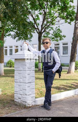 Glücklicher Junge in Schuluniform in einem weißen Hemd, blauer Weste und Sonnenbrille mit Rucksackständern in der Nähe der Schule Stockfoto