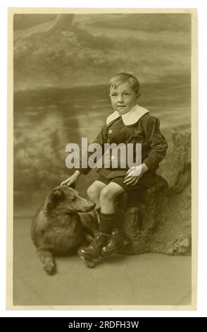 Originell, ungewöhnlich, Studiopostkarte aus der Zeit um WW1, mit einem süßen Jungen, der einen Cordanzug trägt, mit breitem Revers, auch in der Edwardian Times zu sehen, kurze Hosen, gute Stiefel, posieren mit einem makabren, ausgestopften Hundeprop, vom 1916, Großbritannien Stockfoto