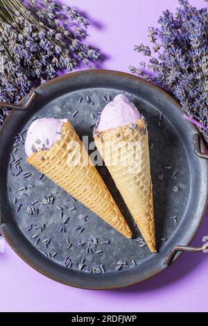 Zwei Lavendeleis in Waffle Cones auf einem Tablett und Blumensträuße aus trockenem Lavendel auf lila Hintergrund. Stockfoto