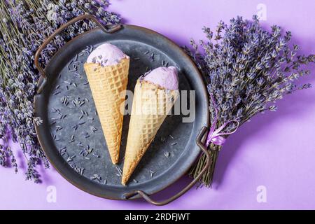 Zwei Lavendeleis in Waffle Cones auf einem Tablett und Blumensträuße aus trockenem Lavendel auf lila Hintergrund. Stockfoto