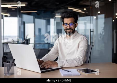 Porträt eines jungen indischen Geschäftsmanns, der im Büro an einem Laptop arbeitet, lächelt und in die Kamera schaut. Stockfoto